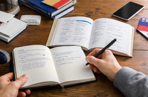 Obraz przedstawia widok z góry na osobę piszącą w otwartym zeszycie w kratkę, trzymającą czarny długopis w prawej dłoni